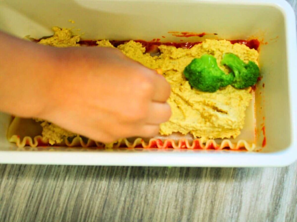 Child’s hand placing broccoli florets on vegan cheese layer while making vegan vegetable lasagna