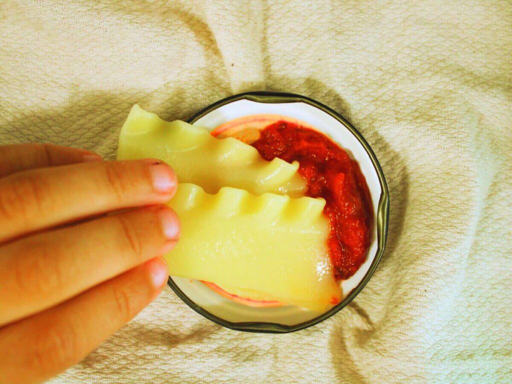 Child’s hand dipping a lasagna noodle into tomato sauce while making vegan vegetable lasagna
