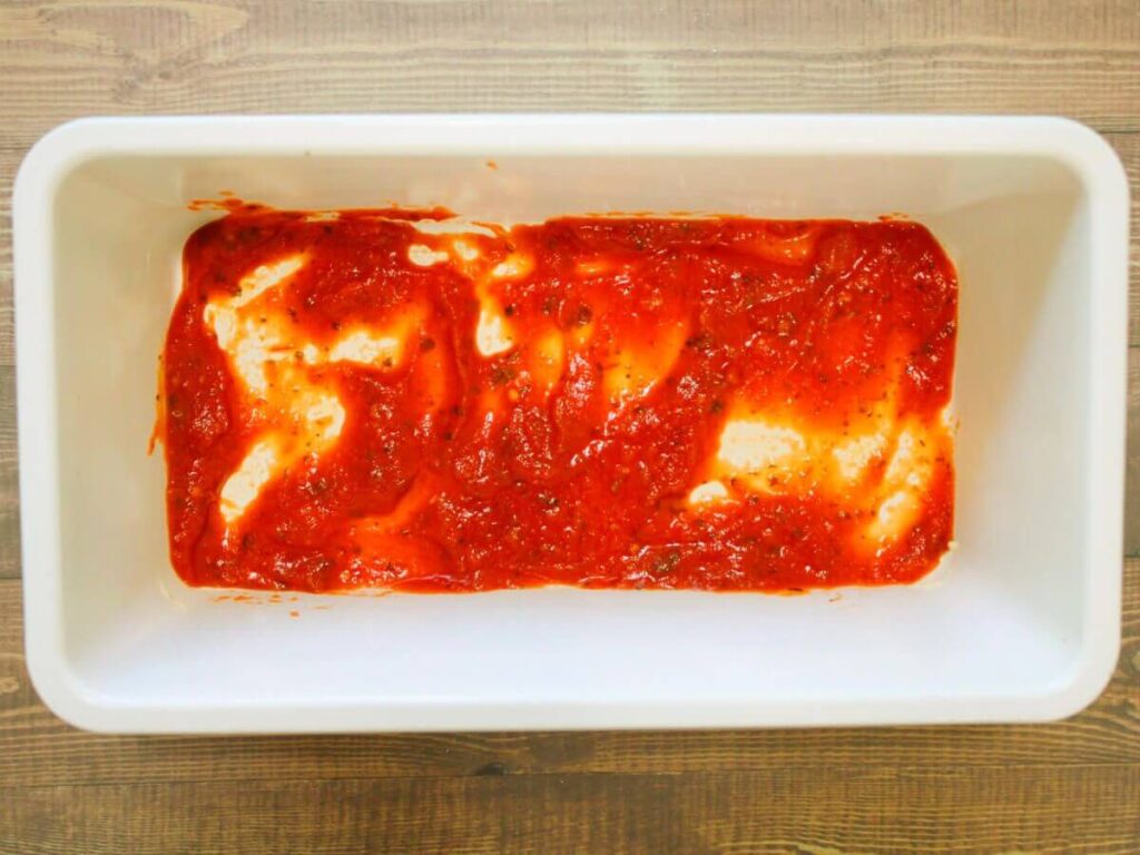 Overhead view of tomato sauce spread in the bottom of a loaf pan for vegan vegetable lasagna