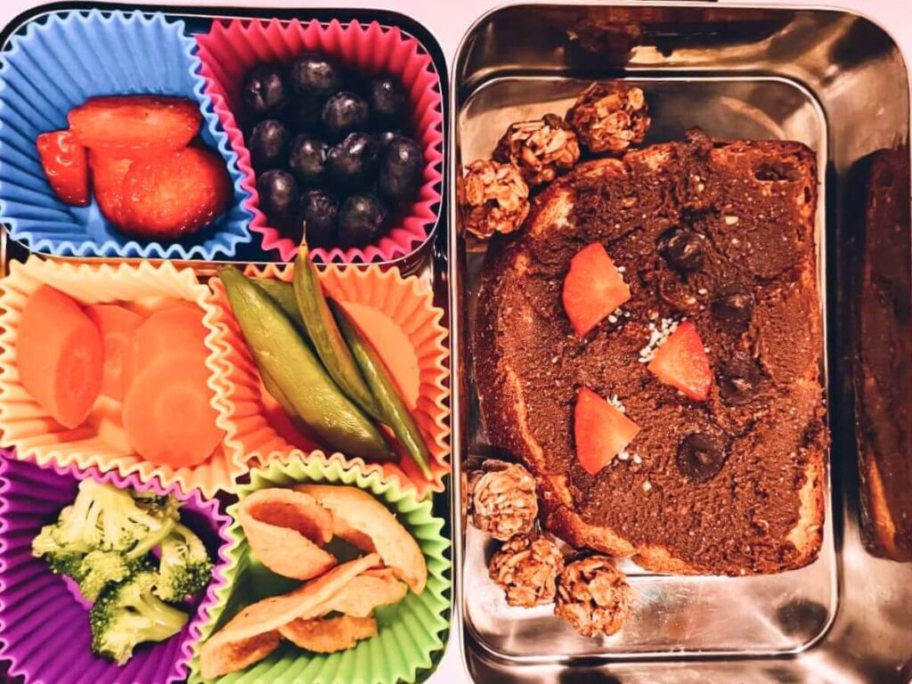 Stainless steel bento lunch with sunflower seed butter sandwich, strawberries, blueberries, broccoli, carrots, snap peas, and tortilla chips.