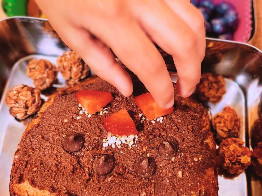 Child placing sliced strawberries on sunflower seed butter sandwich in a stainless steel bento lunchbox.