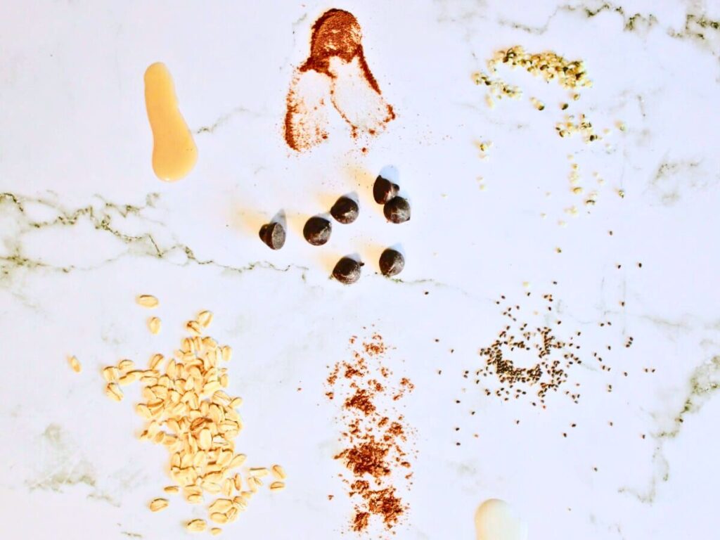 Taste test setup with agave syrup, hemp seeds, cinnamon, cacao powder, oats, chia seeds, and dairy-free chocolate chips on a marble background