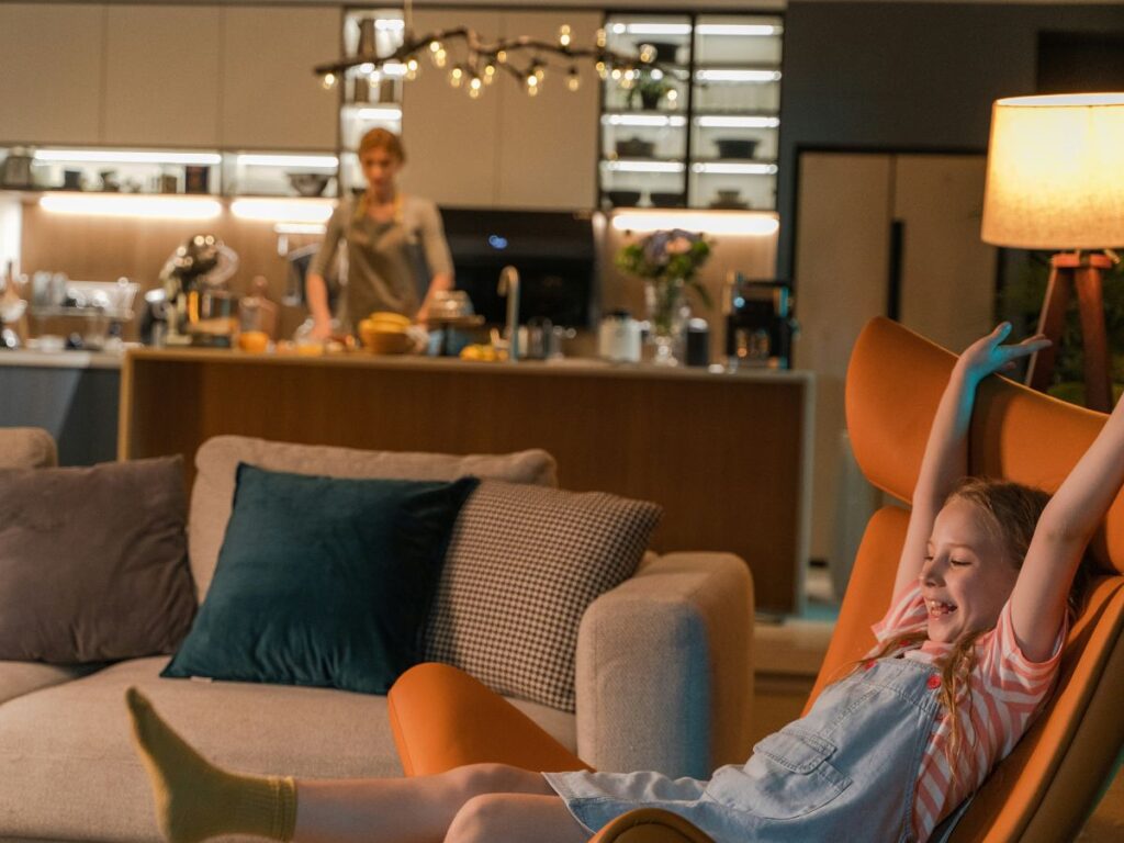 Child relaxing in a living room while a parent cooks nearby, showing connection in a shared space despite different activities